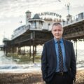 a man in a suit standing on a beach with a pier in the background