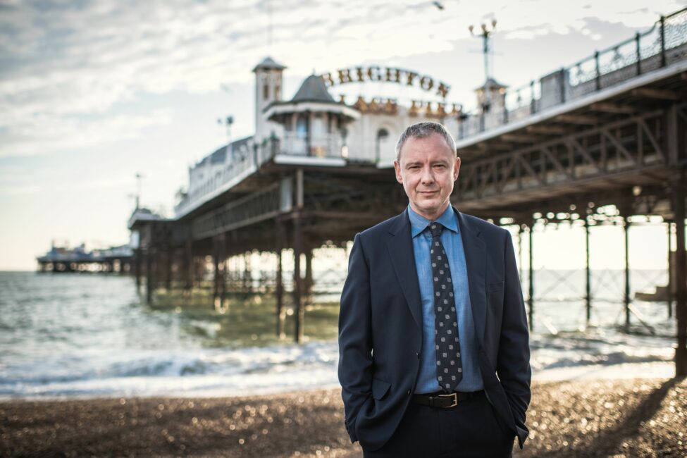 Detective Grace 1 Roy Grace 1 a man in a suit standing on a beach with a pier in the background